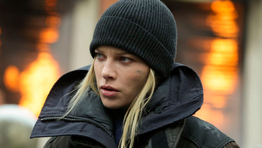 A atriz Lauren German com o uniforme de bombeiros e chamas ao fundo